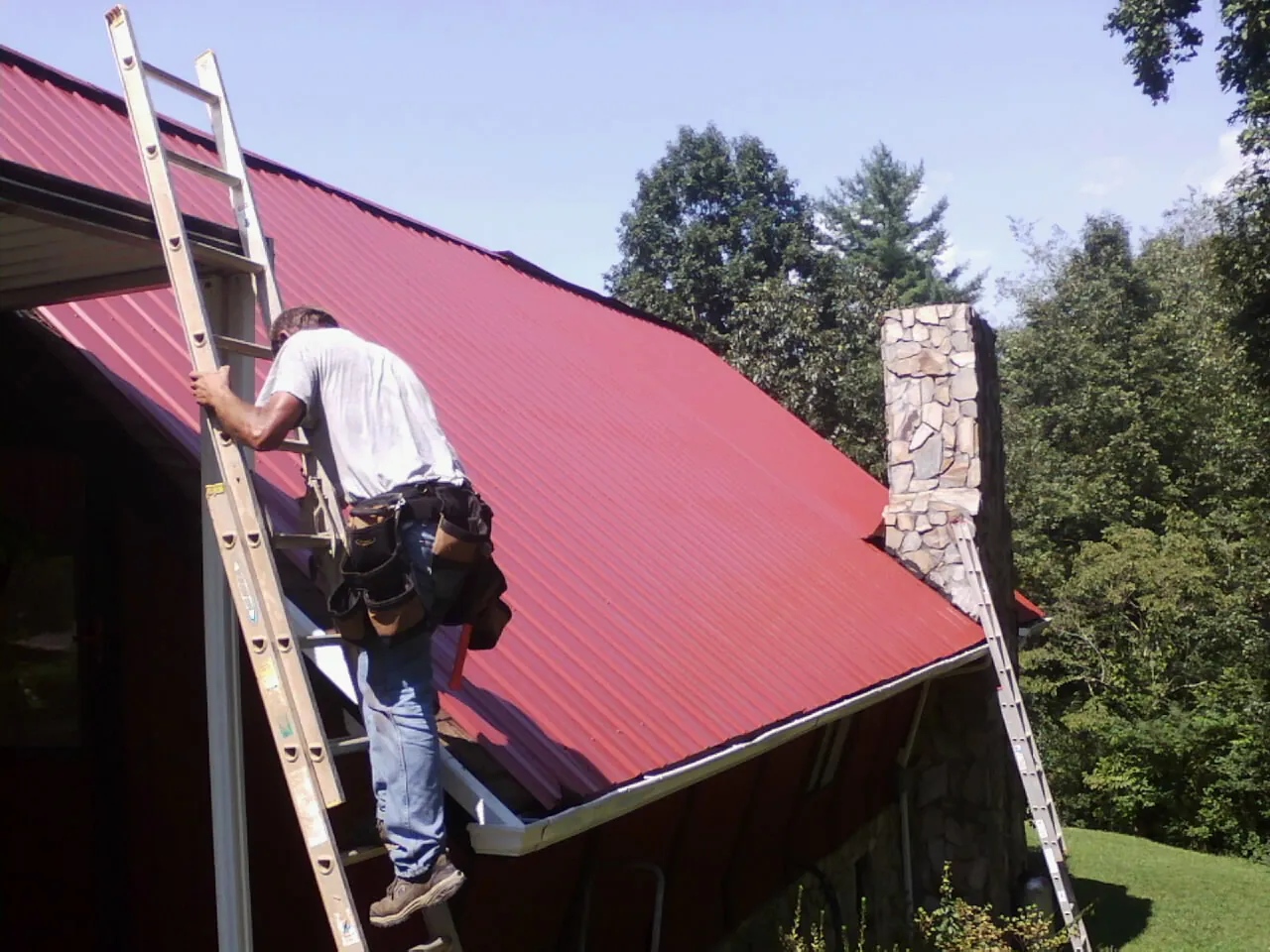 Roofer on a ladder beside a red metal roof and stone chimney — Soffit & Fascia Repair by Roofing Brothers in Loma Linda